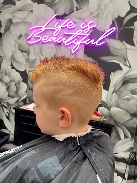 Boy's short textured cut with a faded side, under a Life is Beautiful neon sign